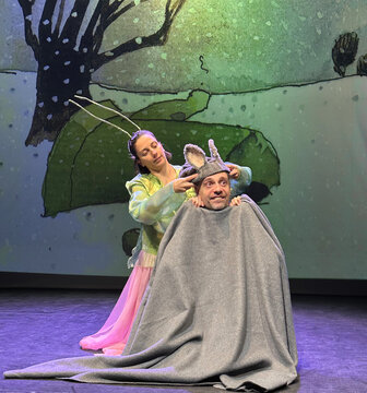 Eine Frau in buntem Kostüm setzt einem Mann mit grauem Decke und Ohr-Accessoire ein Tier-Hausschuhe auf, auf Bühnenhintergrund.