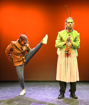 Zwei Bühnenkünstler: Einer in einem braunen Mantel tanzt mit einem Bein in der Luft, der andere hält eine Geige und trägt ein Kleid.