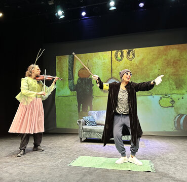 Zwei Schauspieler auf der Bühne: einer mit Violine und Gefieder, der andere gestikulierend vor einer projizierten Kulisse.