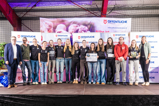 Gruppenfoto einer Sportmannschaft mit Trainer, alle in einheitlichen Shirts, auf einer Bühne während einer Veranstaltung.