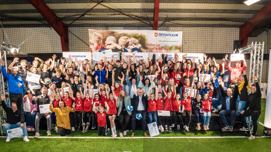 Gruppenszene in einer Sporthalle mit vielen Menschen in Sportkleidung, die jubelnd ihre Hände heben und Urkunden präsentieren.