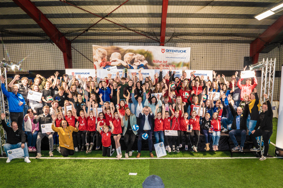Gruppenszene in einer Sporthalle mit vielen Menschen in Sportkleidung, die jubelnd ihre Hände heben und Urkunden präsentieren.