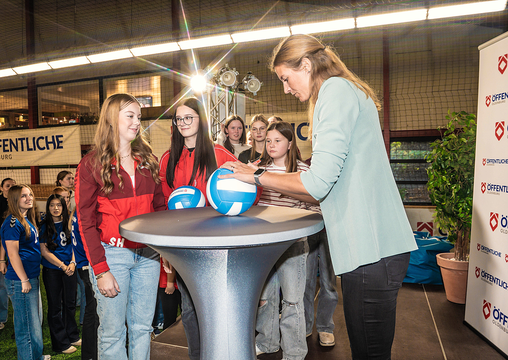 Eine Sportlerin signiert Bälle an einem Stehtisch, umgeben von jungen Fans in einer Sporthalle.