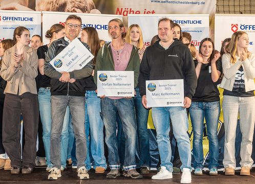 Drei Männer auf einer Bühne halten Auszeichnungen für die besten Trainer, umgeben von applaudierenden Frauen.