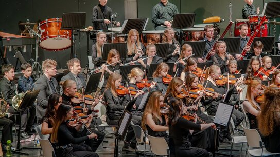Jugendliches Orchester auf der Bühne, Spieler mit verschiedenen Instrumenten in schwarzer Kleidung, konzentriert beim Musizieren.