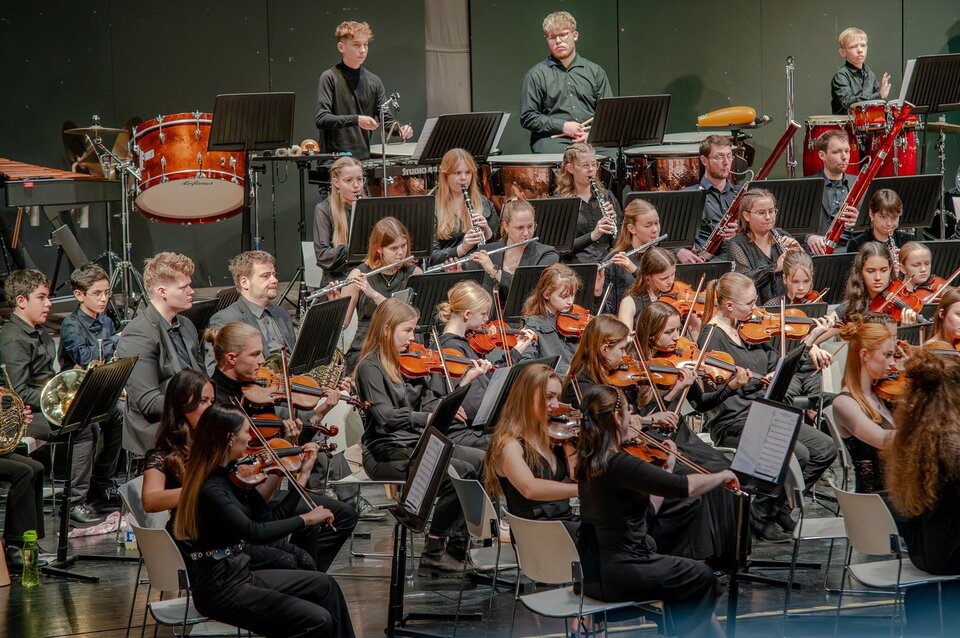 Jugendliches Orchester auf der Bühne, Spieler mit verschiedenen Instrumenten in schwarzer Kleidung, konzentriert beim Musizieren.
