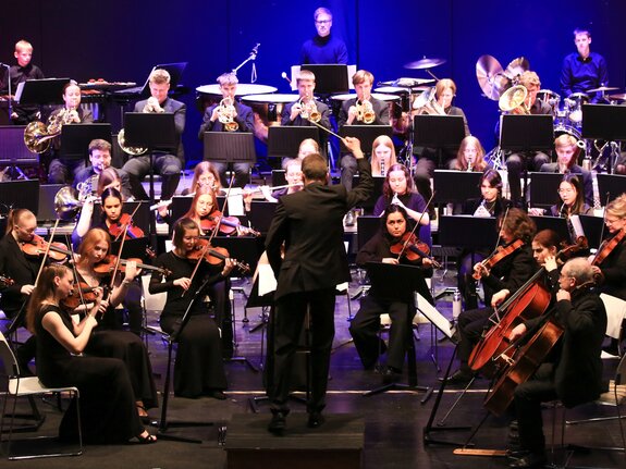 Ein großes Orchester in formeller Kleidung spielt auf einer Bühne, dirigiert von einem Dirigenten vor zahlreichen Musikern.