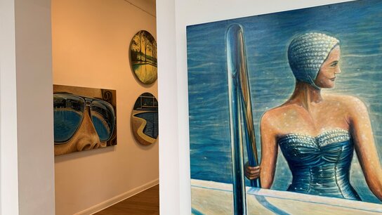 In einer Galerie sind zwei Wände mit verschiedenen Kunstwerken dekoriert, darunter ein Porträt einer Schwimmerin.