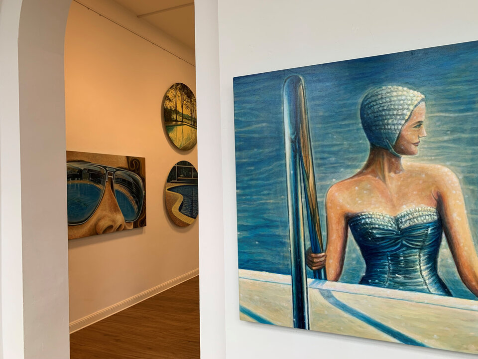 In einer Galerie sind zwei Wände mit verschiedenen Kunstwerken dekoriert, darunter ein Porträt einer Schwimmerin.