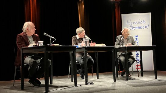 Klaus Modick, Felicitas Hoppe und Bernd Eilert sitzen an Tischen in einem Veranstaltungsraum, umgeben von Scheinwerfern und einem Willkommensbanner.
