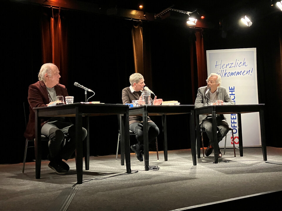 Klaus Modick, Felicitas Hoppe und Bernd Eilert sitzen an Tischen in einem Veranstaltungsraum, umgeben von Scheinwerfern und einem Willkommensbanner.