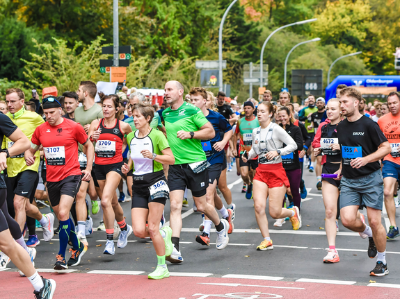 Läuferinnen und Läufer in bunter Sportkleidung starten einen Wettlauf auf einer Straßenstrecke. Bäume im Hintergrund.