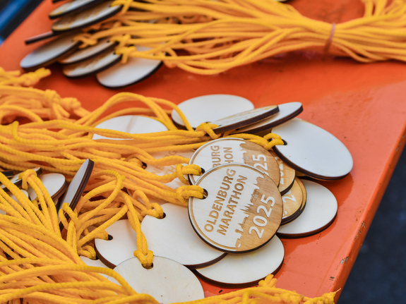 Medaille mit gelben Bändern für den Oldenburger Marathon 2025, auf einer orangefarbenen Fläche angeordnet.