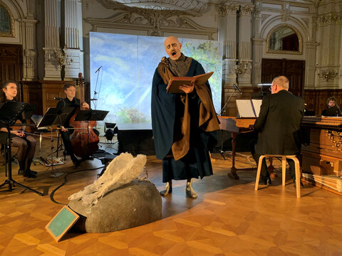 Ein Sänger in einem eleganten Outfit performt auf einer Bühne, umgeben von Musikern und einem Klavier.