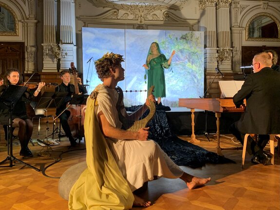 Szenische Darbietung mit Musiker*innen in eleganter Kleidung, Sänger*in in grünem Gewand und Darsteller mit Laute auf einem Holzfußboden.