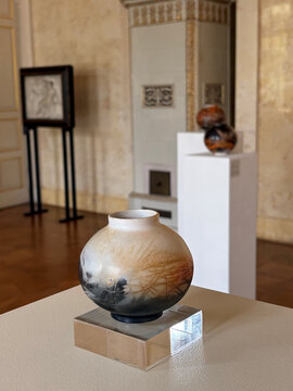 Bunte Vase mit runder Form und kunstvollem Muster, auf einem glatten Standfuß in einem lichtdurchfluteten Raum.