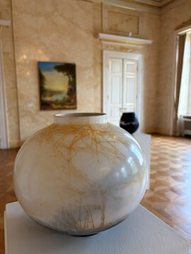 Runde, cremefarbene Vase mit feinen goldenen Linien, im Hintergrund eine Kunstgalerie mit einem Gemälde und weiteren Objekten.