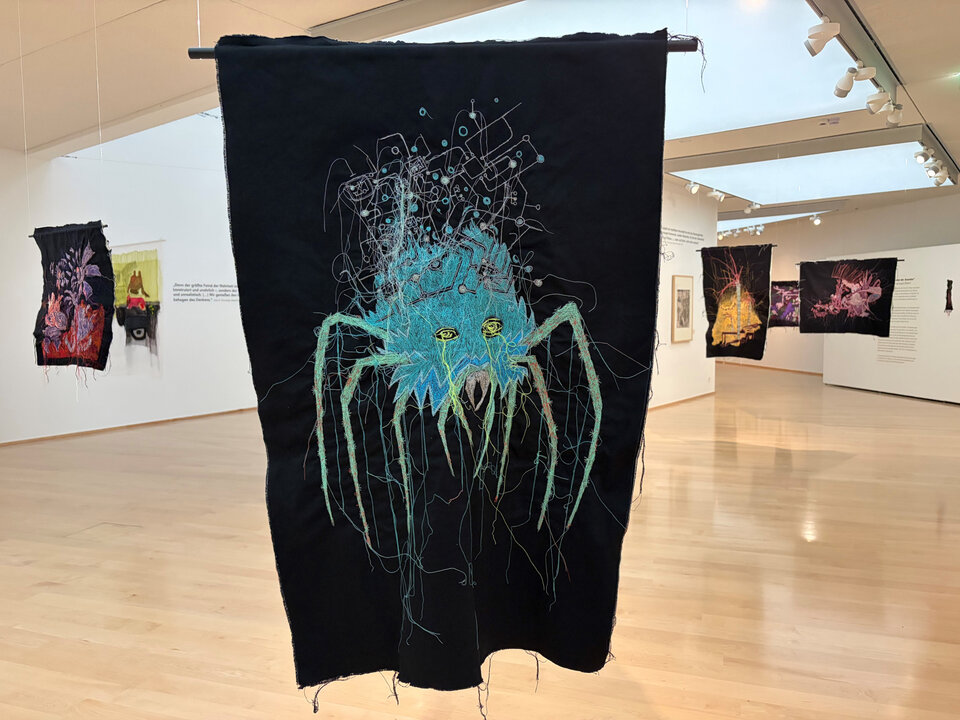 Schwarzes Textil mit einem blauen Spinnenmotiv und farbigen, verstreuten Fäden, im Hintergrund mehrere weitere Textilien.