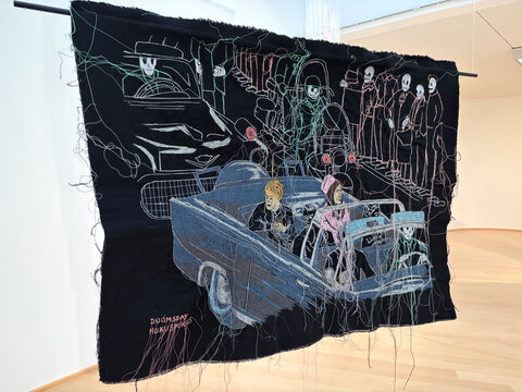 Buntgestickte Wandbehängung zeigt eine Autoscene mit Figuren, umgeben von schematischen, gespenstischen Silhouetten.