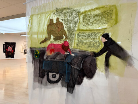 Kunstinstallation mit schematischen Figuren und Schrift auf einem durchsichtigen Stoffhang. Hintergrund zeigt weitere Kunstwerke.