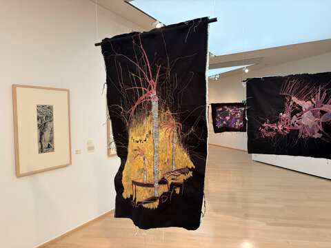 Hängendes Textilkunstwerk mit bunten Stickereien auf schwarzem Hintergrund in einer Galerie.