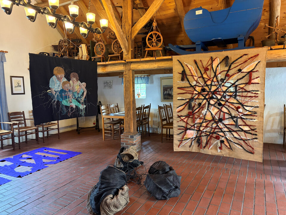 Kunstwerke aus Textilien hängen in einem rustikalen Raum mit Holzbalken, Stühlen und einem blauen Gefährt.
