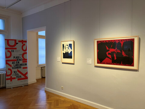 Ausstellungsraum mit zwei Bildern an der Wand und einem Plakat auf der linken Seite, welches für eine Kunstausstellung wirbt.