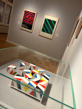 Skulpturen in Glasvitrine, umgeben von bunten geometrischen Kunstwerken an der Wand in einer Galerie.