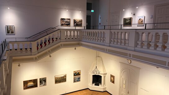 Blick auf eine Galerie mit Kunstwerken an den Wänden und einem offenen Treppenbereich im Vordergrund.