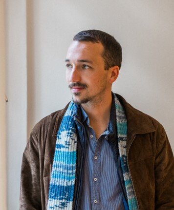 Mann mit braunem Jacket und blauen, gestreiften Schal blickt nachdenklich zur Seite in einem hellen Raum.