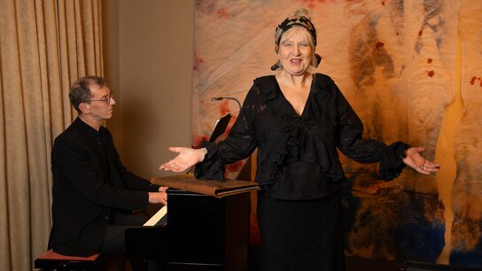 Sängerin in schwarzer Kleidung tritt auf, während ein Pianist am Klavier begleitet. Bunte Wandgestaltung im Hintergrund.