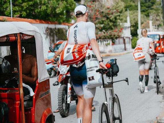 Sportler mit Fahrrad trägt eine Sporttasche, während weitere Teilnehmer auf einer Straße vorbeifahren.
