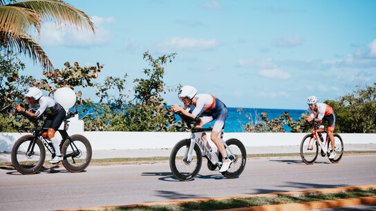 Drei Sportler in Radrennausrüstung fahren auf einer Küstenstraße, hinter ihnen der Ozean und Palmen.
