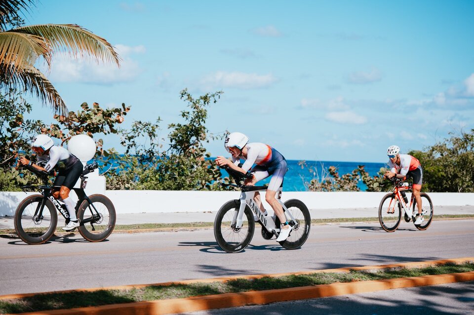 Drei Sportler in Radrennausrüstung fahren auf einer Küstenstraße, hinter ihnen der Ozean und Palmen.