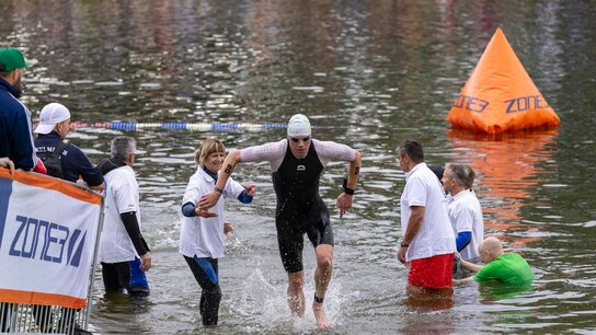 Ein Schwimmer verlässt das Wasser, während Helfer ihn unterstützen. Im Hintergrund ist eine Wendemarke sichtbar.