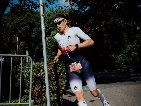 Ein männlicher Sportler läuft in Wettkleidung auf einer Straße, vorbei an einem Straßenschild mit der Aufschrift "Jadeallee".