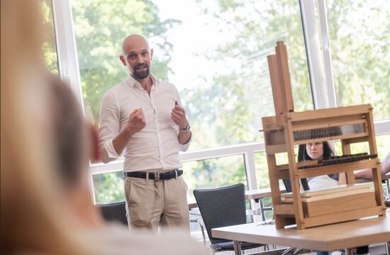 Mann in weißem Hemd hält Vortrag vor Sitzenden, im Hintergrund ein Webstuhl und große Fenster mit Blick ins Freie.