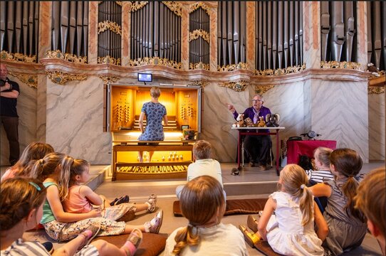 Eine Gruppe von Kindern sitzt auf dem Boden und schaut einem Musiker an einer Orgel zu, während er ihnen etwas erklärt.