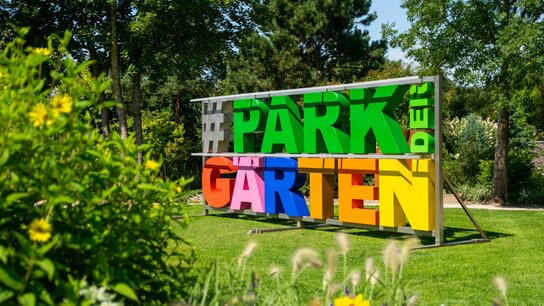 Bunte Buchstaben bilden den Schriftzug "#PARK der GARTEN" vor einer grünen Kulisse mit Blumen.
