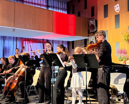 Ensemble spielt auf der Bühne, Musiker mit verschiedenen Instrumenten in formeller Kleidung, freundliche Atmosphäre im Hintergrund.