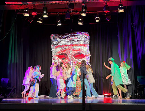 Farbenfrohe Theateraufführung mit mehreren Schauspielern, die sich um eine große, silberne Maske auf der Bühne versammeln.