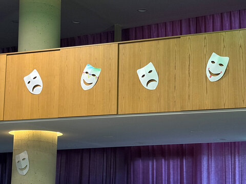 Weißes Holzregal mit vier stilisierten Masken (zwei lachend, zwei traurig) an einer Wand, im Hintergrund lila Vorhänge.