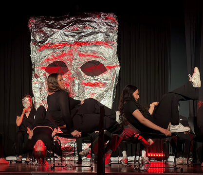 Tanzperformance mit mehreren Tänzern in schwarzer Kleidung vor einer großen, silbernen Maske auf der Bühne.