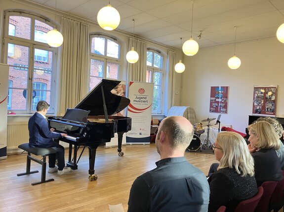 Ein junger Künstler sitzt am Klavier in einem Veranstaltungsraum. Zuschauer gucken ihm zu. Im Hintergrund ist ein Rollup zui Jugend musiziert zu sehen.