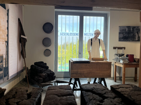 Eine historische Bäckerei mit einem Mannequin in weißer Kleidung, einem Holztisch und gestapeltem Brot im Vordergrund.