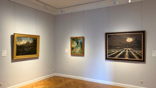 Drei Gemälde in einer Galerie, eines mit Landschaft, eines mit abstrakten Formen und das dritte mit einer dramatischen Himmelsszene.
