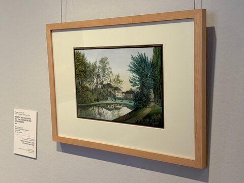 Landschaftsbild mit Häusern und Bäumen, reflektierender Teich, in einem schlichten Holzrahmen.
