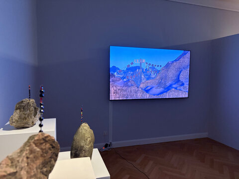 Moderne Kunstinstallation mit einer Wand, auf der ein Bildschirm Landschaftsvisualisierungen zeigt, und Steinskulpturen im Vordergrund.