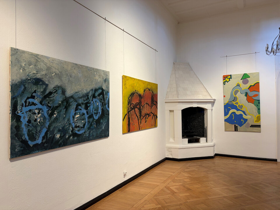 Wände einer Galerie mit abstrakten Gemälden in verschiedenen Farben; ein Kamin steht in der Ecke.