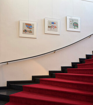Rote Treppe mit schwarzem Geländer, an der Wand hängen drei gerahmte Kunstwerke.